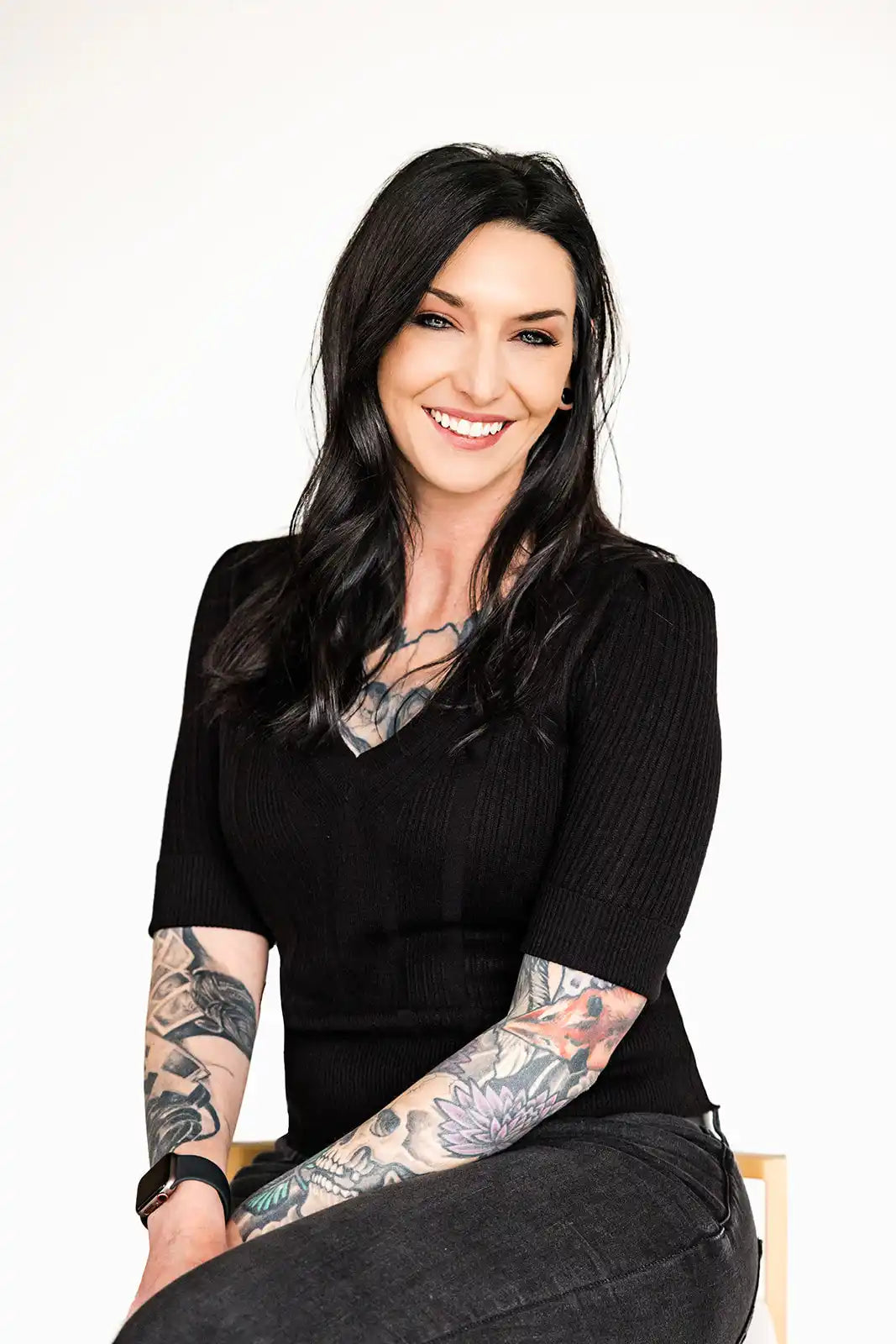 A smiling woman in black clothing with visible arm tattoos.